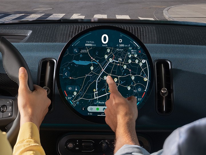 MINI Électromobilité – écran OLED central avec navigation. Conducteur et passager interagissant avec l’écran d’infodivertissement circulaire MINI affichant la navigation.